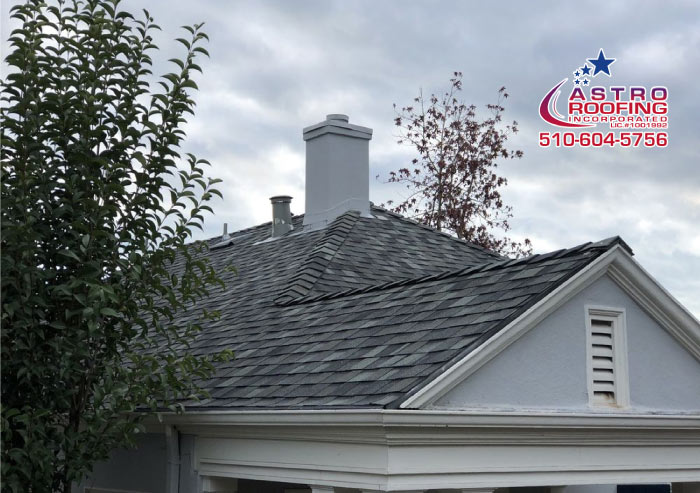 Close-up of a dark grey shingle roof and a chimney, featuring the Astro Roofing logo and phone number