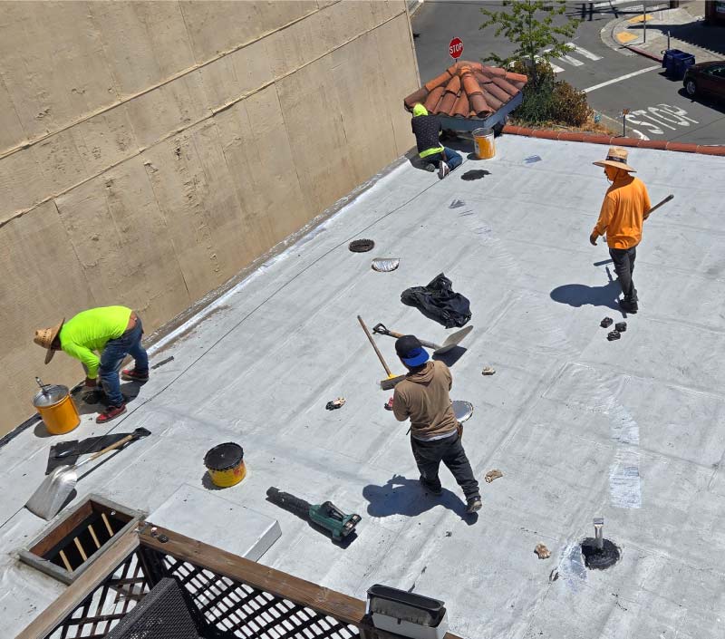 Astro Roofing team performing flat commercial roofing maintenance and applying a protective coating to a commercial roof system near a street corner in the Bay Area.